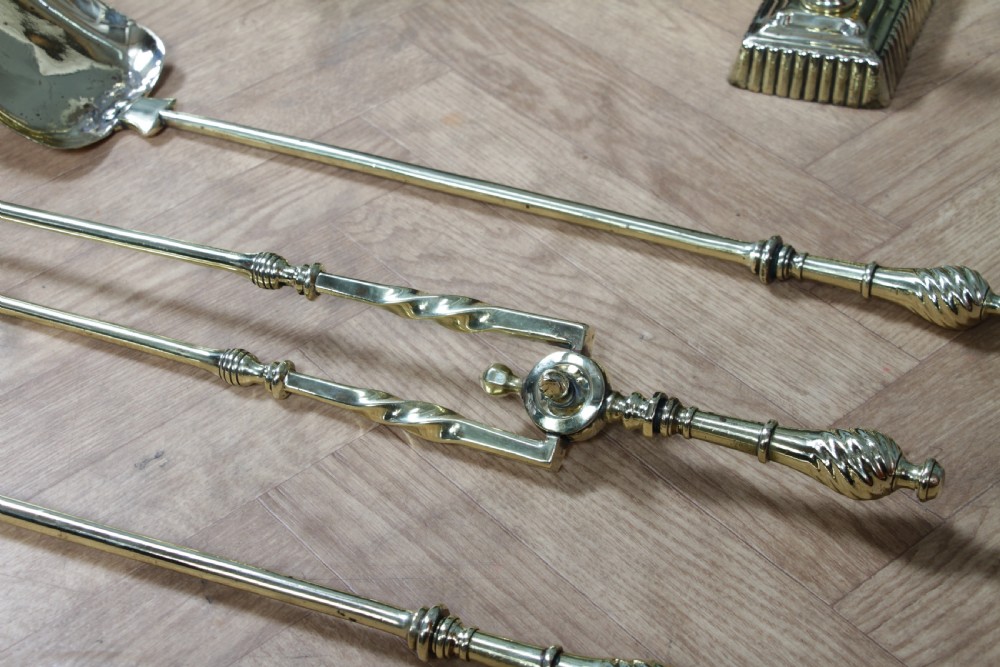 A late Victorian polished brass companion set featuring a shovel, tongs, and poker supported horizontally across two urn-topped brass firedogs. View X