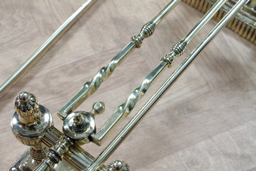 A late Victorian polished brass companion set featuring a shovel, tongs, and poker supported horizontally across two urn-topped brass firedogs. View XII