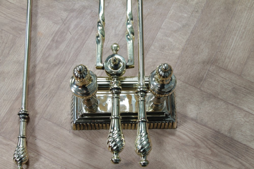 A late Victorian polished brass companion set featuring a shovel, tongs, and poker supported horizontally across two urn-topped brass firedogs. View XIII
