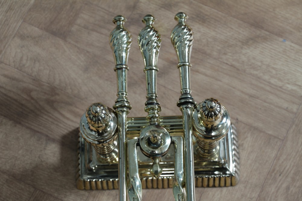 A late Victorian polished brass companion set featuring a shovel, tongs, and poker supported horizontally across two urn-topped brass firedogs. View XV