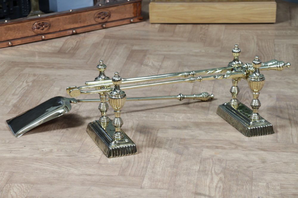 A late Victorian polished brass companion set featuring a shovel, tongs, and poker supported horizontally across two urn-topped brass firedogs. View XVII