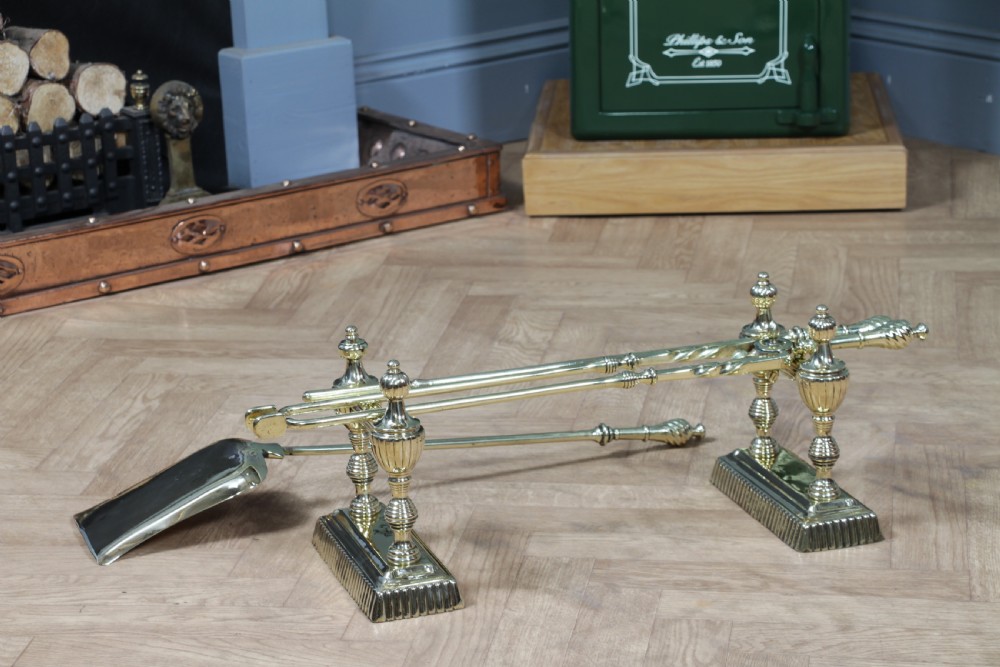 A late Victorian polished brass companion set featuring a shovel, tongs, and poker supported horizontally across two urn-topped brass firedogs. View XVIII