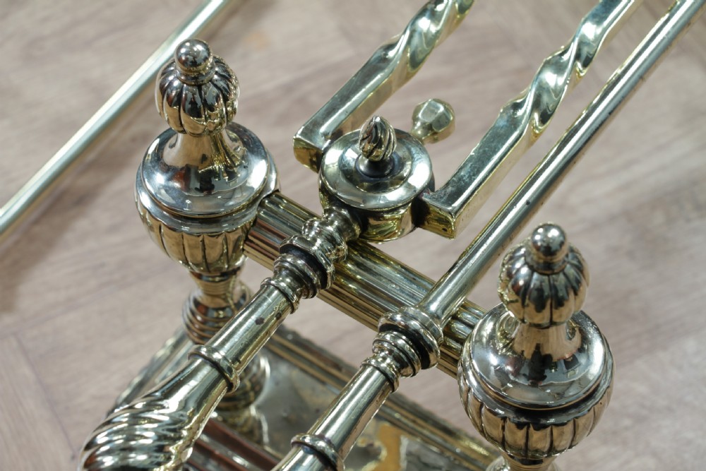 A late Victorian polished brass companion set featuring a shovel, tongs, and poker supported horizontally across two urn-topped brass firedogs. View XX
