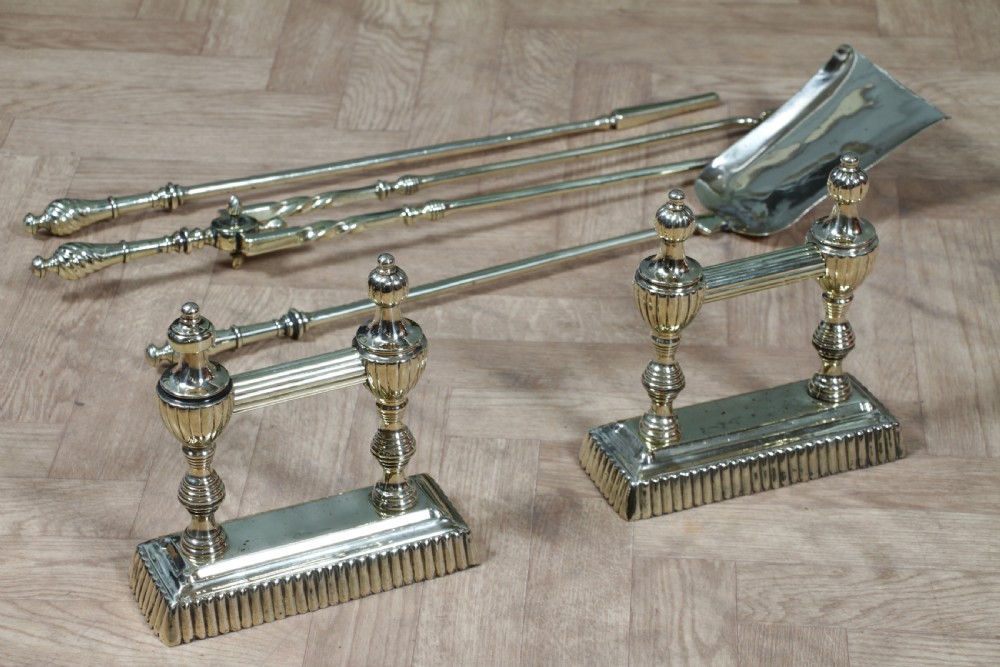 A late Victorian polished brass companion set featuring a shovel, tongs, and poker supported horizontally across two urn-topped brass firedogs. View XXI
