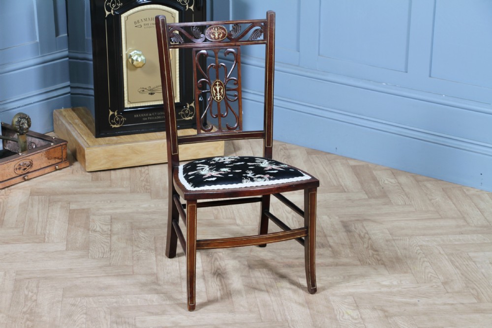 An Edwardian mahogany nursing chair featuring an inlaid pierced back splat, decorative stringing, and a black silk upholstered seat with embroidered floral motifs.