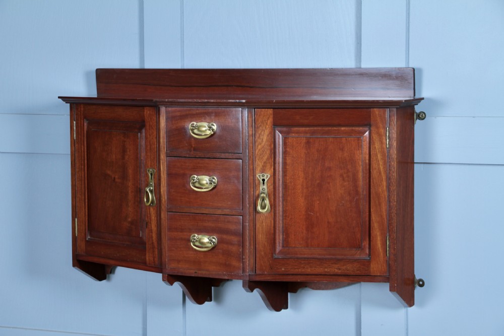 A small Edwardian mahogany wall hanging cabinet featuring two outer cupboards, three central drawers, brass handles, and a low back gallery.