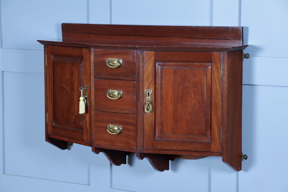 A small Edwardian mahogany wall hanging cabinet featuring two outer cupboards, three central drawers, brass handles, and a low back gallery. View II