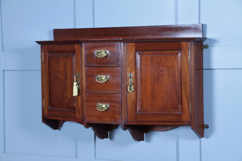 A small Edwardian mahogany wall hanging cabinet featuring two outer cupboards, three central drawers, brass handles, and a low back gallery. View III