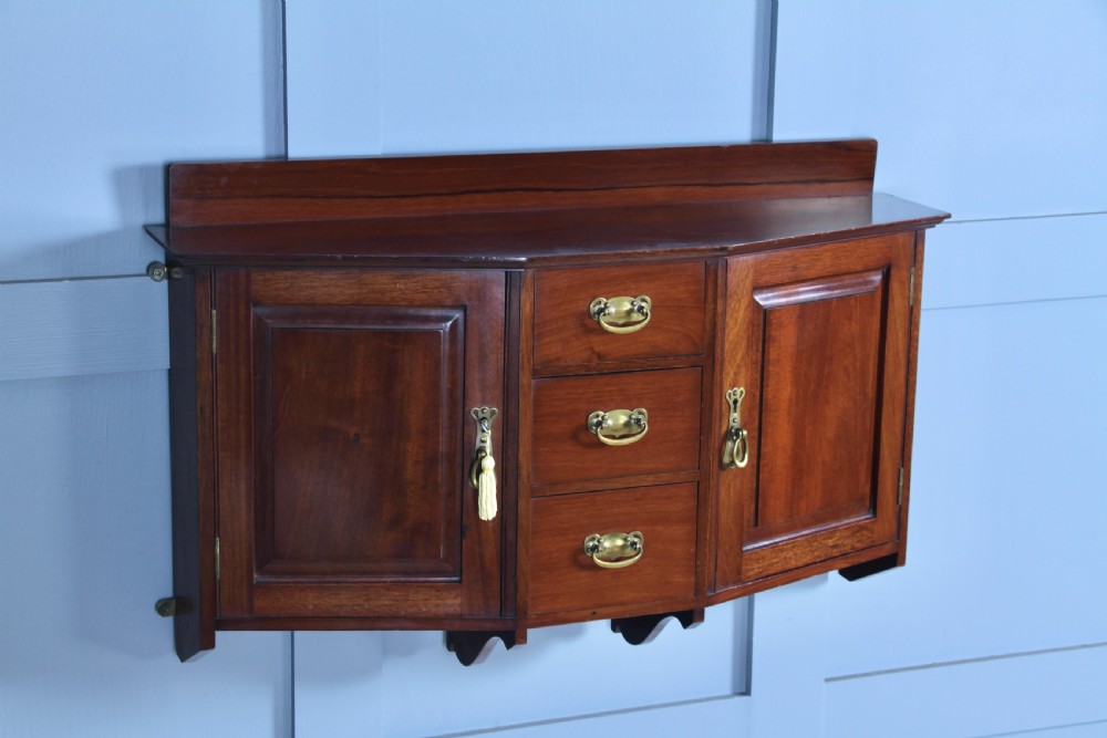 A small Edwardian mahogany wall hanging cabinet featuring two outer cupboards, three central drawers, brass handles, and a low back gallery. View V