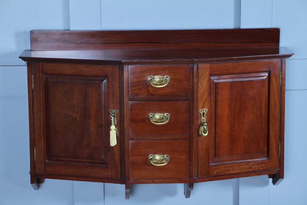 A small Edwardian mahogany wall hanging cabinet featuring two outer cupboards, three central drawers, brass handles, and a low back gallery. View VI