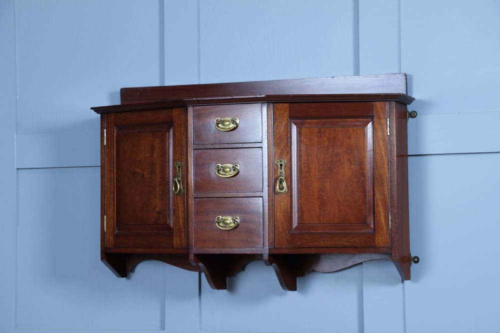 A small Edwardian mahogany wall hanging cabinet featuring two outer cupboards, three central drawers, brass handles, and a low back gallery. View VII