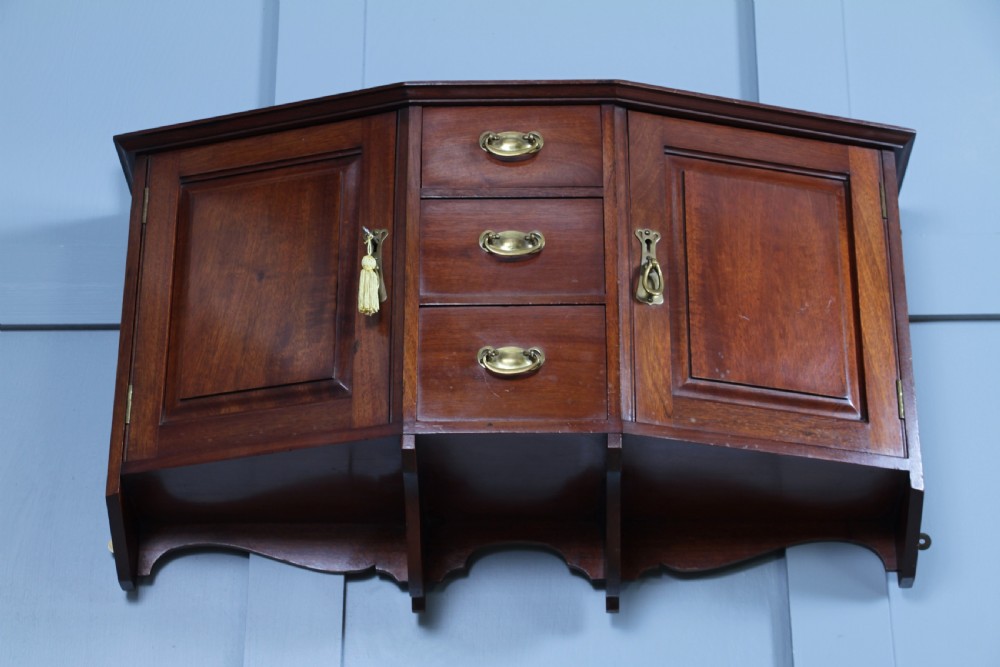 A small Edwardian mahogany wall hanging cabinet featuring two outer cupboards, three central drawers, brass handles, and a low back gallery. View VIII
