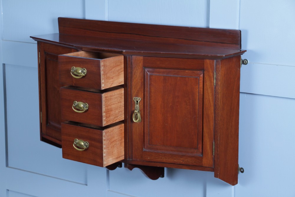 A small Edwardian mahogany wall hanging cabinet featuring two outer cupboards, three central drawers, brass handles, and a low back gallery. View XV