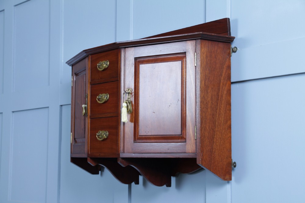 A small Edwardian mahogany wall hanging cabinet featuring two outer cupboards, three central drawers, brass handles, and a low back gallery. View XVIII