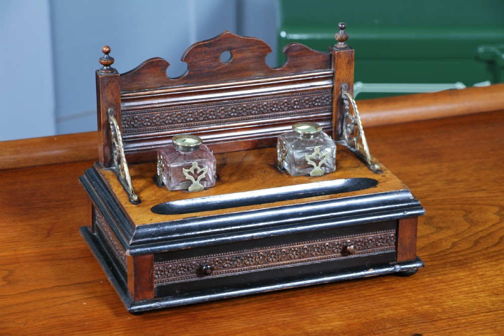 An Edwardian wooden inkstand featuring two glass inkwells, a shaped back gallery with finials, brass side supports, a pen groove, and a lower drawer.