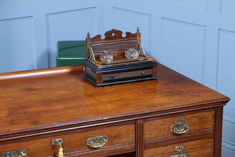 An Edwardian wooden inkstand featuring two glass inkwells, a shaped back gallery with finials, brass side supports, a pen groove, and a lower drawer. View II