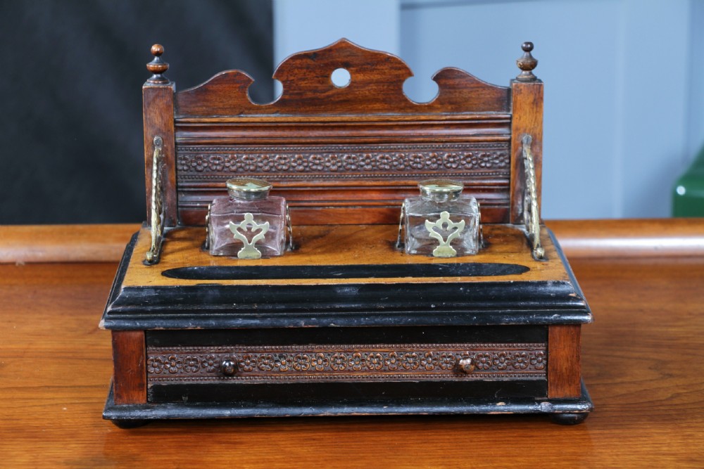 An Edwardian wooden inkstand featuring two glass inkwells, a shaped back gallery with finials, brass side supports, a pen groove, and a lower drawer. View III