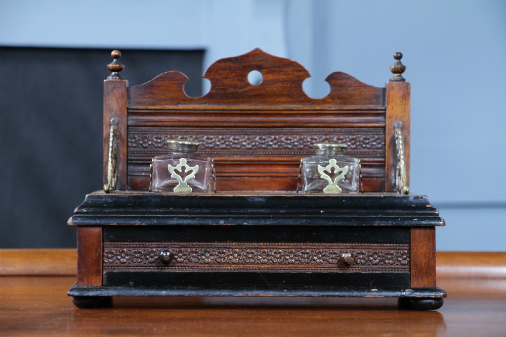 An Edwardian wooden inkstand featuring two glass inkwells, a shaped back gallery with finials, brass side supports, a pen groove, and a lower drawer. View V