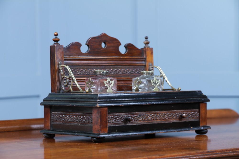 An Edwardian wooden inkstand featuring two glass inkwells, a shaped back gallery with finials, brass side supports, a pen groove, and a lower drawer. View VI