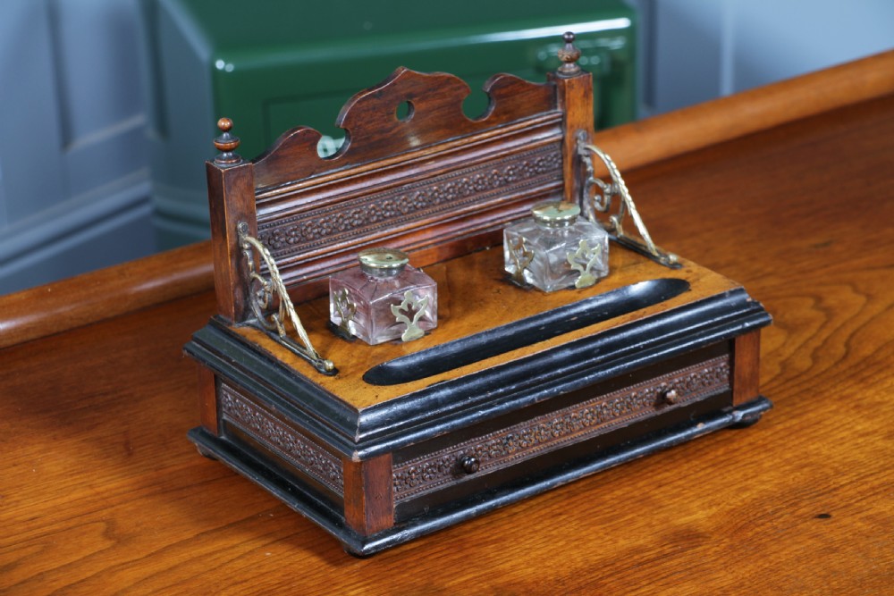 An Edwardian wooden inkstand featuring two glass inkwells, a shaped back gallery with finials, brass side supports, a pen groove, and a lower drawer. View VII