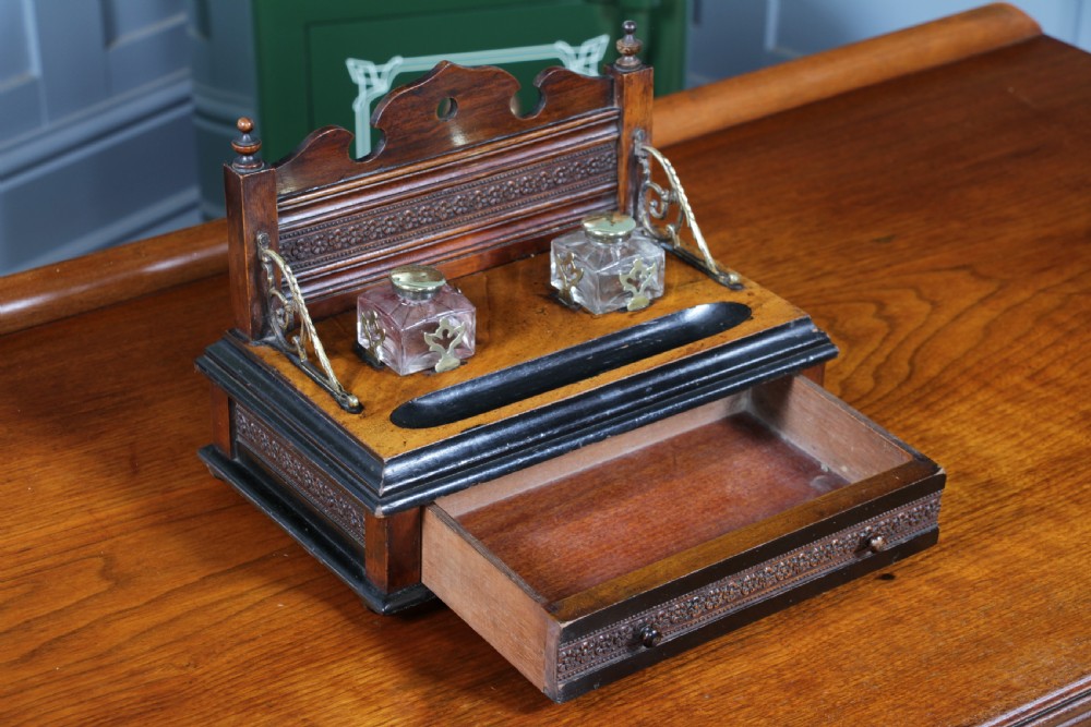 An Edwardian wooden inkstand featuring two glass inkwells, a shaped back gallery with finials, brass side supports, a pen groove, and a lower drawer. View VIII