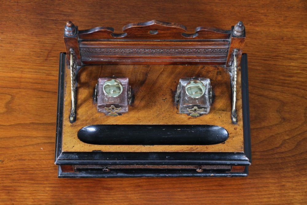 An Edwardian wooden inkstand featuring two glass inkwells, a shaped back gallery with finials, brass side supports, a pen groove, and a lower drawer. View X