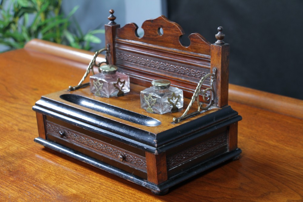 An Edwardian wooden inkstand featuring two glass inkwells, a shaped back gallery with finials, brass side supports, a pen groove, and a lower drawer. View XI