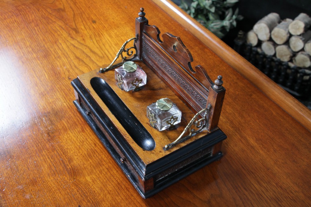An Edwardian wooden inkstand featuring two glass inkwells, a shaped back gallery with finials, brass side supports, a pen groove, and a lower drawer. View XII