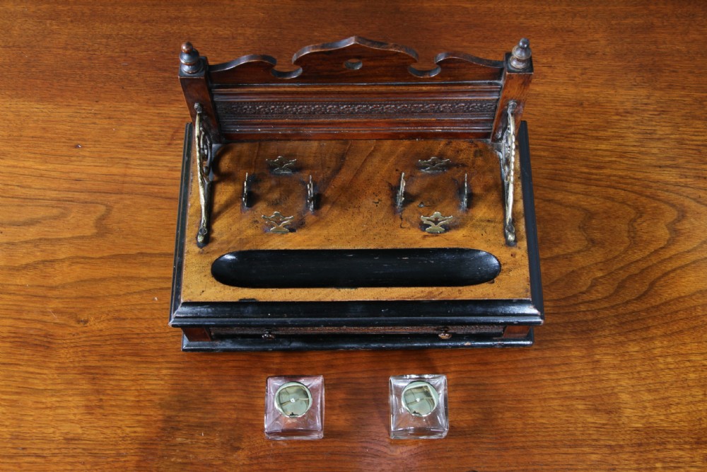 An Edwardian wooden inkstand featuring two glass inkwells, a shaped back gallery with finials, brass side supports, a pen groove, and a lower drawer. View XIV