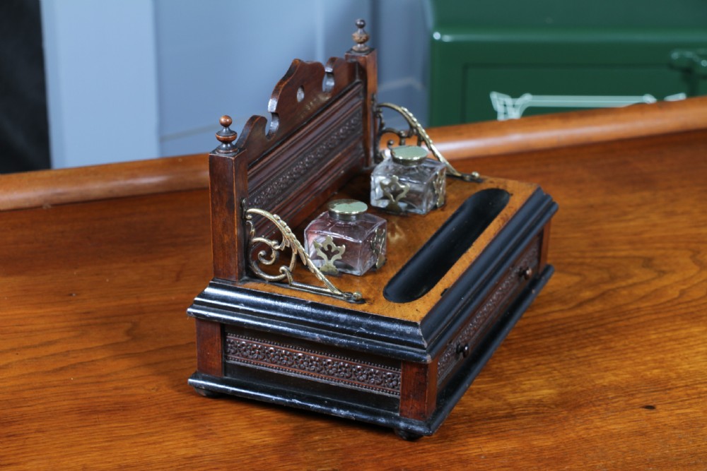 An Edwardian wooden inkstand featuring two glass inkwells, a shaped back gallery with finials, brass side supports, a pen groove, and a lower drawer. View XVII
