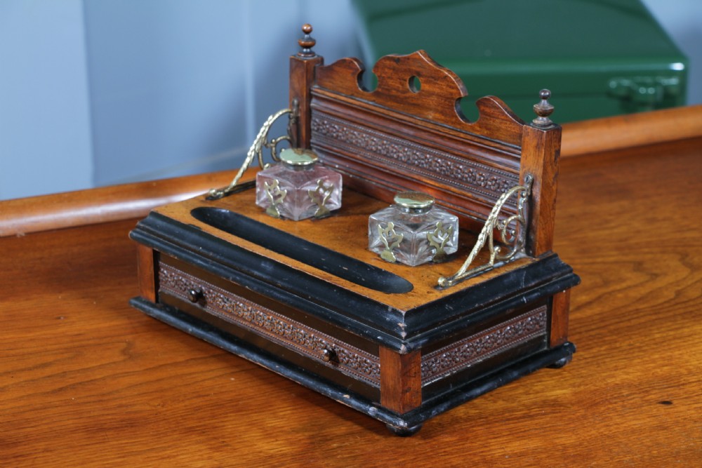 An Edwardian wooden inkstand featuring two glass inkwells, a shaped back gallery with finials, brass side supports, a pen groove, and a lower drawer. View XVIII