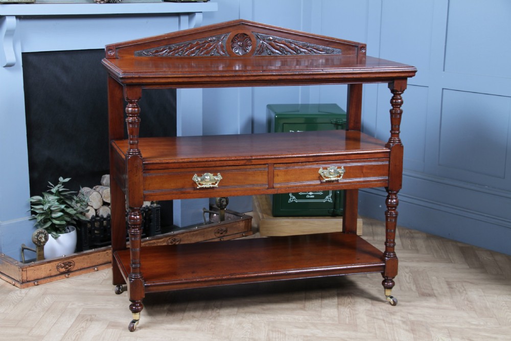 A three-tier Victorian walnut dumb waiter featuring turned columns, carved pediment gallery, two central drawers with brass handles, and brass castors.