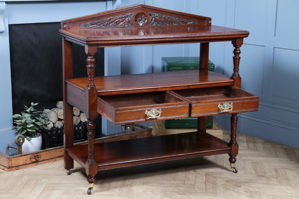 A three-tier Victorian walnut dumb waiter featuring turned columns, carved pediment gallery, two central drawers with brass handles, and brass castors. View II