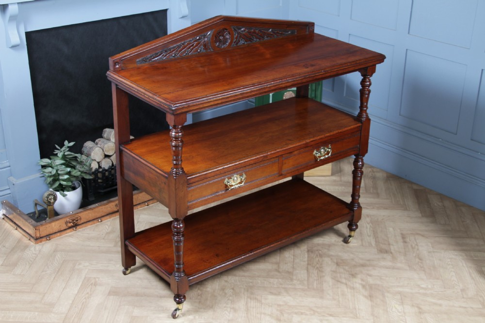 A three-tier Victorian walnut dumb waiter featuring turned columns, carved pediment gallery, two central drawers with brass handles, and brass castors. View III
