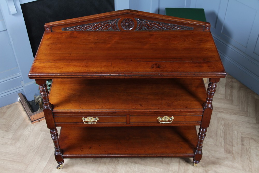 A three-tier Victorian walnut dumb waiter featuring turned columns, carved pediment gallery, two central drawers with brass handles, and brass castors. View V