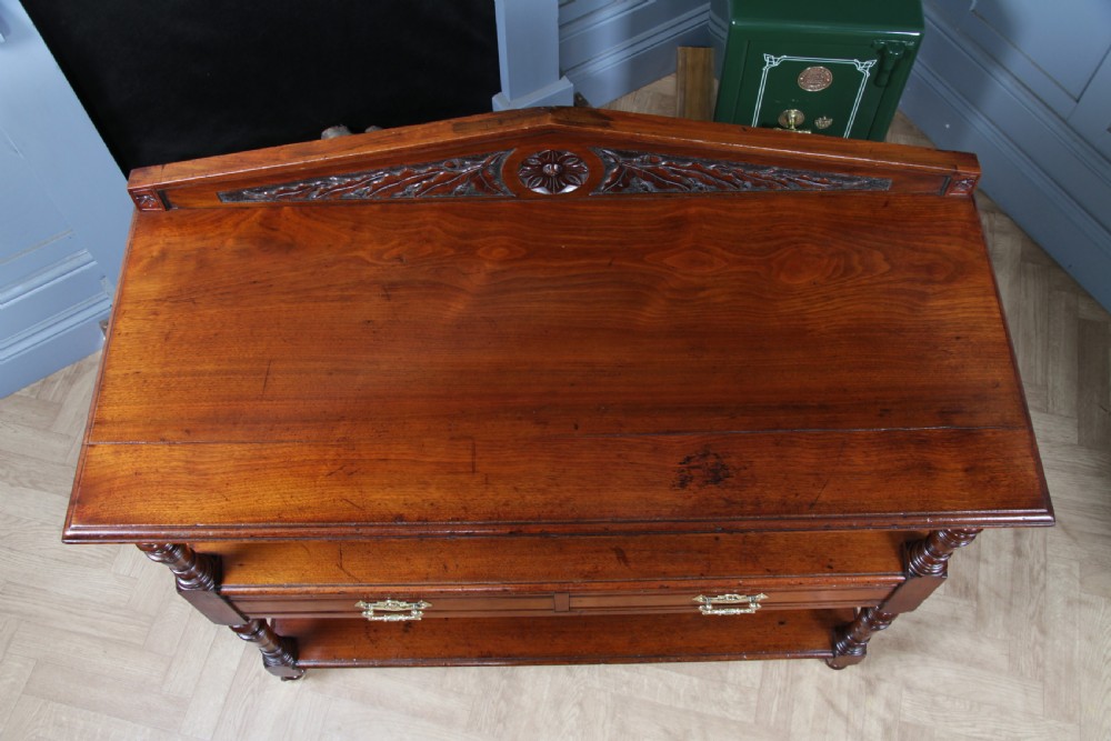 A three-tier Victorian walnut dumb waiter featuring turned columns, carved pediment gallery, two central drawers with brass handles, and brass castors. View X