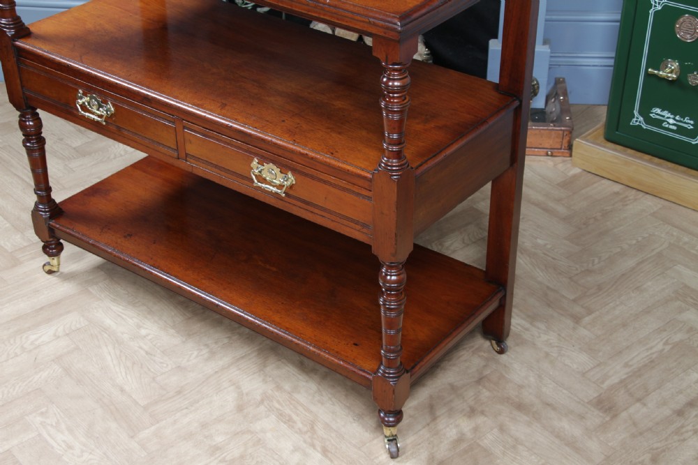 A three-tier Victorian walnut dumb waiter featuring turned columns, carved pediment gallery, two central drawers with brass handles, and brass castors. View XI