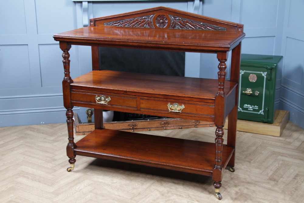 A three-tier Victorian walnut dumb waiter featuring turned columns, carved pediment gallery, two central drawers with brass handles, and brass castors. View XII