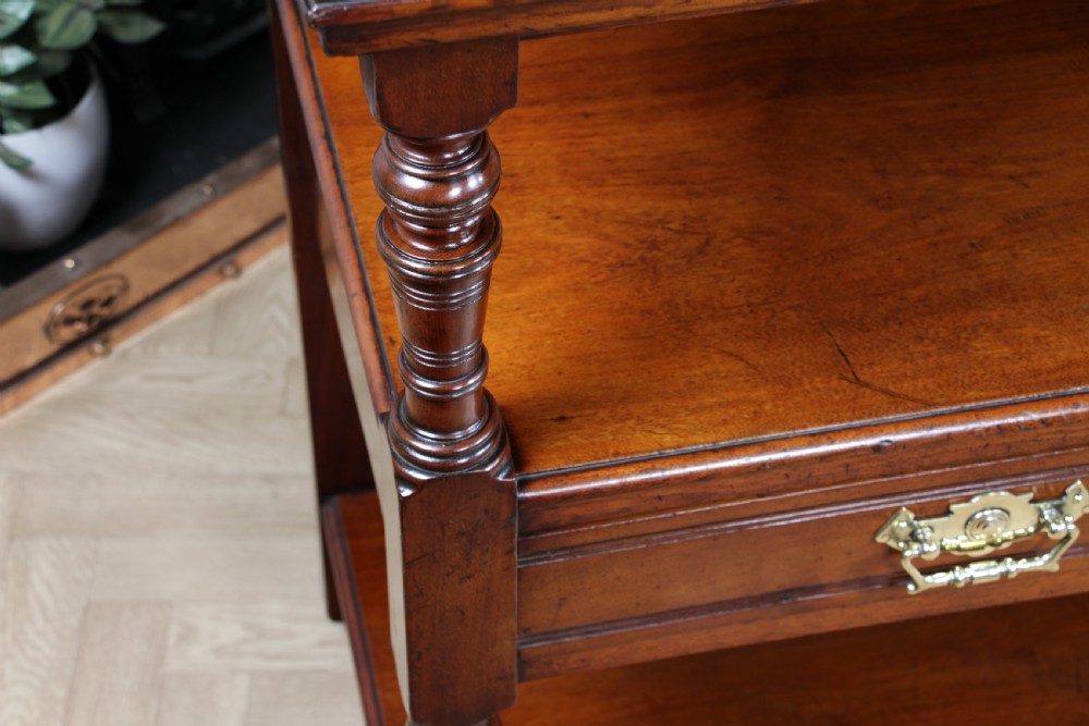 A three-tier Victorian walnut dumb waiter featuring turned columns, carved pediment gallery, two central drawers with brass handles, and brass castors. View XV