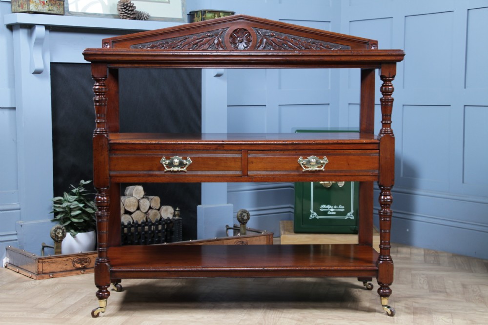 A three-tier Victorian walnut dumb waiter featuring turned columns, carved pediment gallery, two central drawers with brass handles, and brass castors. View XVI