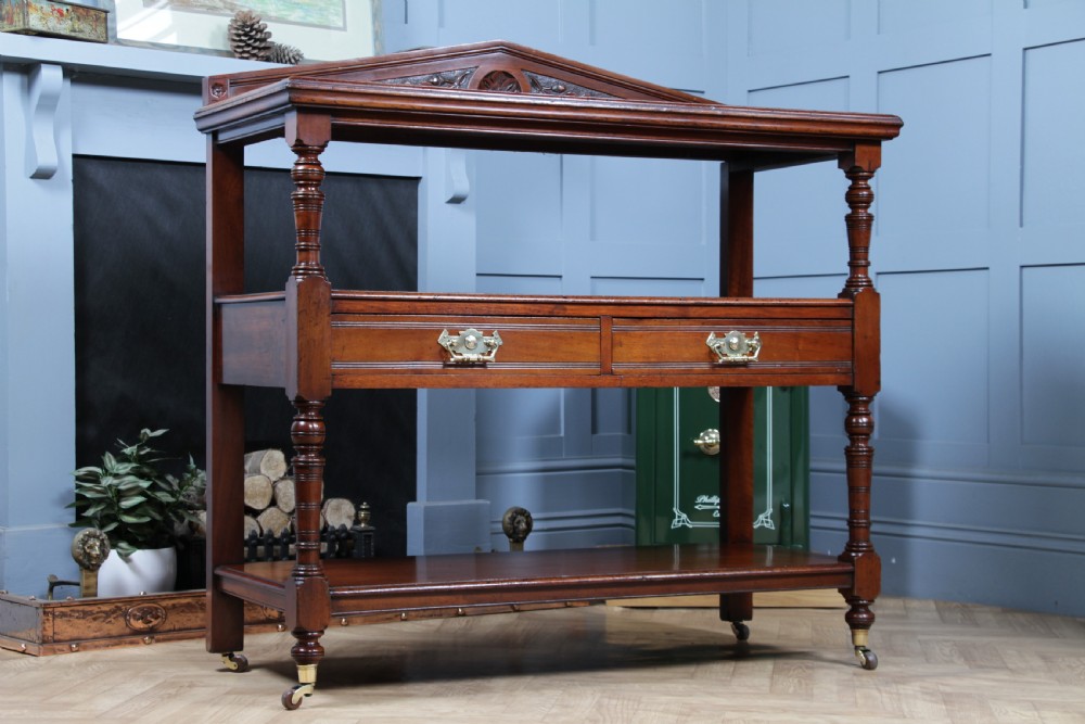A three-tier Victorian walnut dumb waiter featuring turned columns, carved pediment gallery, two central drawers with brass handles, and brass castors. View XVII
