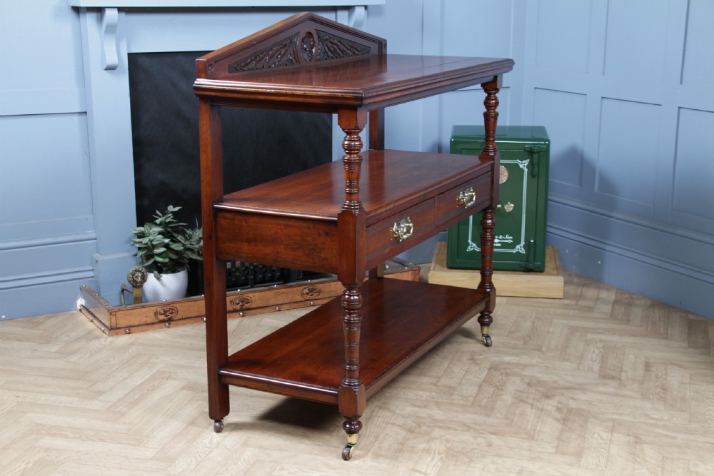 A three-tier Victorian walnut dumb waiter featuring turned columns, carved pediment gallery, two central drawers with brass handles, and brass castors. View XXI