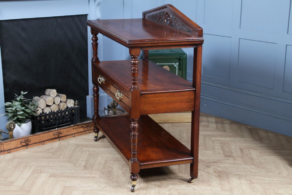 A three-tier Victorian walnut dumb waiter featuring turned columns, carved pediment gallery, two central drawers with brass handles, and brass castors. View XXII