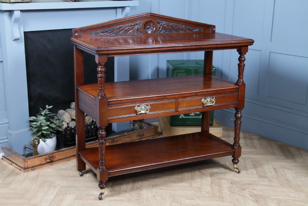 A three-tier Victorian walnut dumb waiter featuring turned columns, carved pediment gallery, two central drawers with brass handles, and brass castors. View XXIV