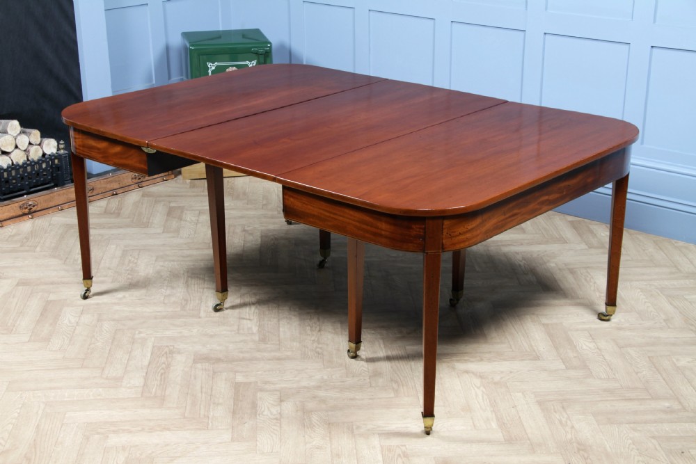 An extended Georgian mahogany dining table featuring semi-circular D-ends, a central drop-leaf section, square tapered legs, and brass castors.
