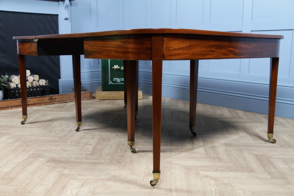 An extended Georgian mahogany dining table featuring semi-circular D-ends, a central drop-leaf section, square tapered legs, and brass castors. View VI