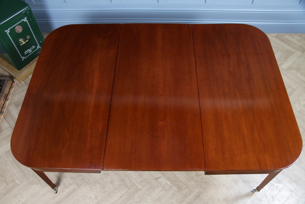 An extended Georgian mahogany dining table featuring semi-circular D-ends, a central drop-leaf section, square tapered legs, and brass castors. View VII