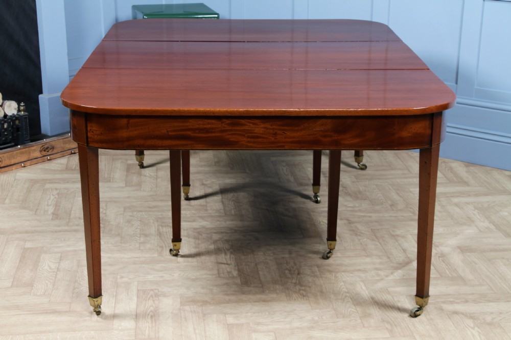An extended Georgian mahogany dining table featuring semi-circular D-ends, a central drop-leaf section, square tapered legs, and brass castors. View XIX