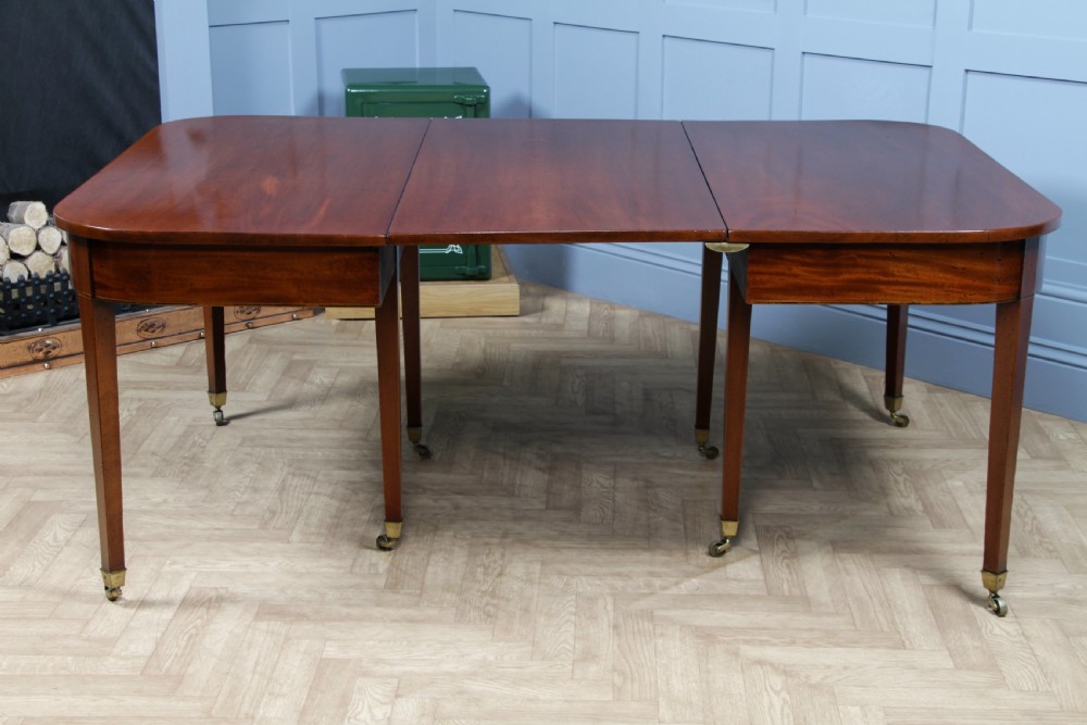 An extended Georgian mahogany dining table featuring semi-circular D-ends, a central drop-leaf section, square tapered legs, and brass castors. View XX