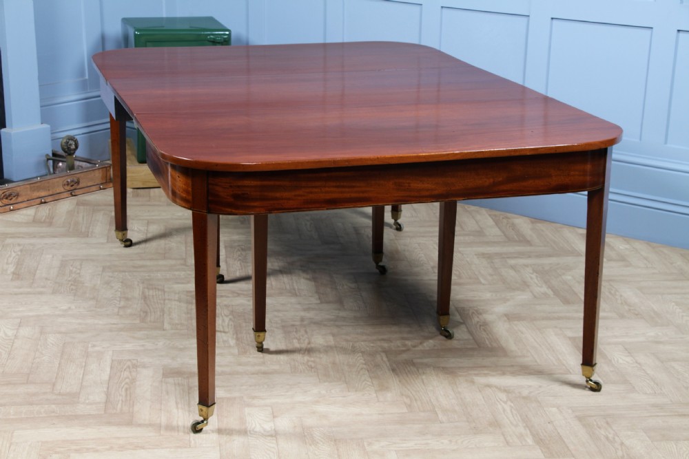 An extended Georgian mahogany dining table featuring semi-circular D-ends, a central drop-leaf section, square tapered legs, and brass castors. View XXI